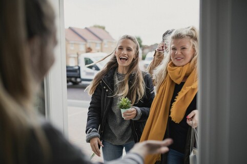 three people arive at the open door of a home, greeted by the host. They are smiling and carrying small gifts.